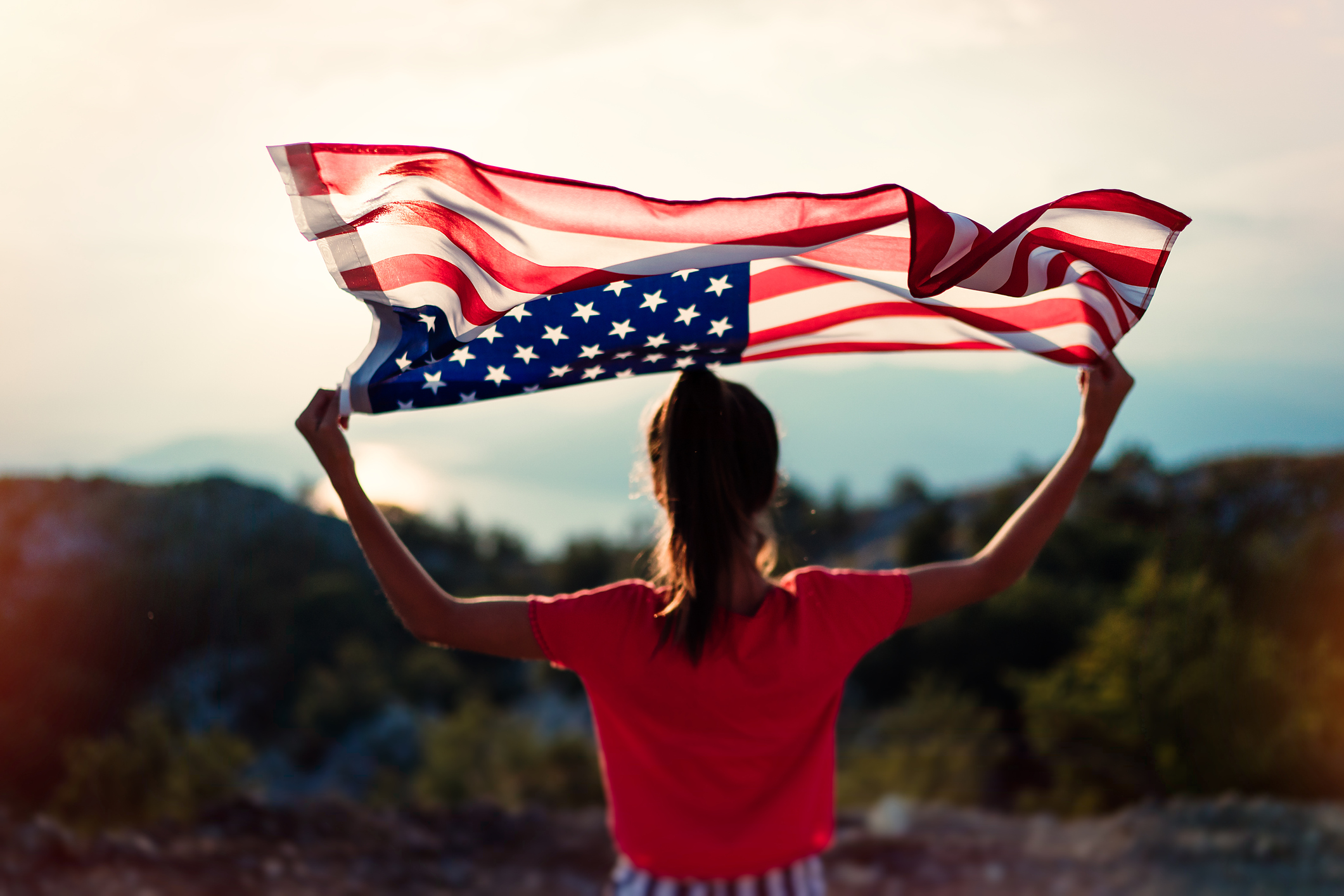 Teenager girl  is waving American flag on top of mountain at sky background