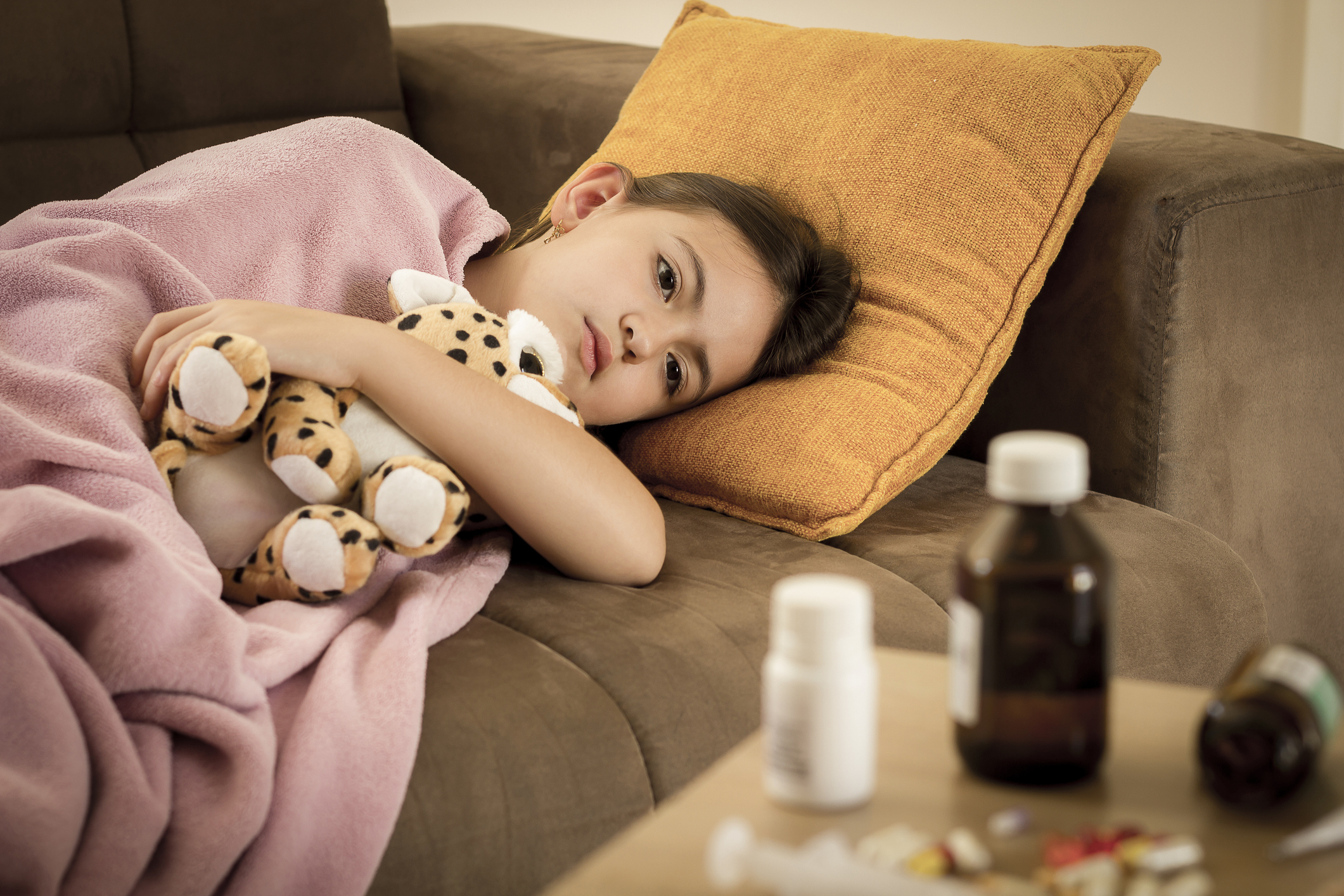 Sick little girl covered in blanket is hugging teddy bear and looking sadly on medicine while lying on couch