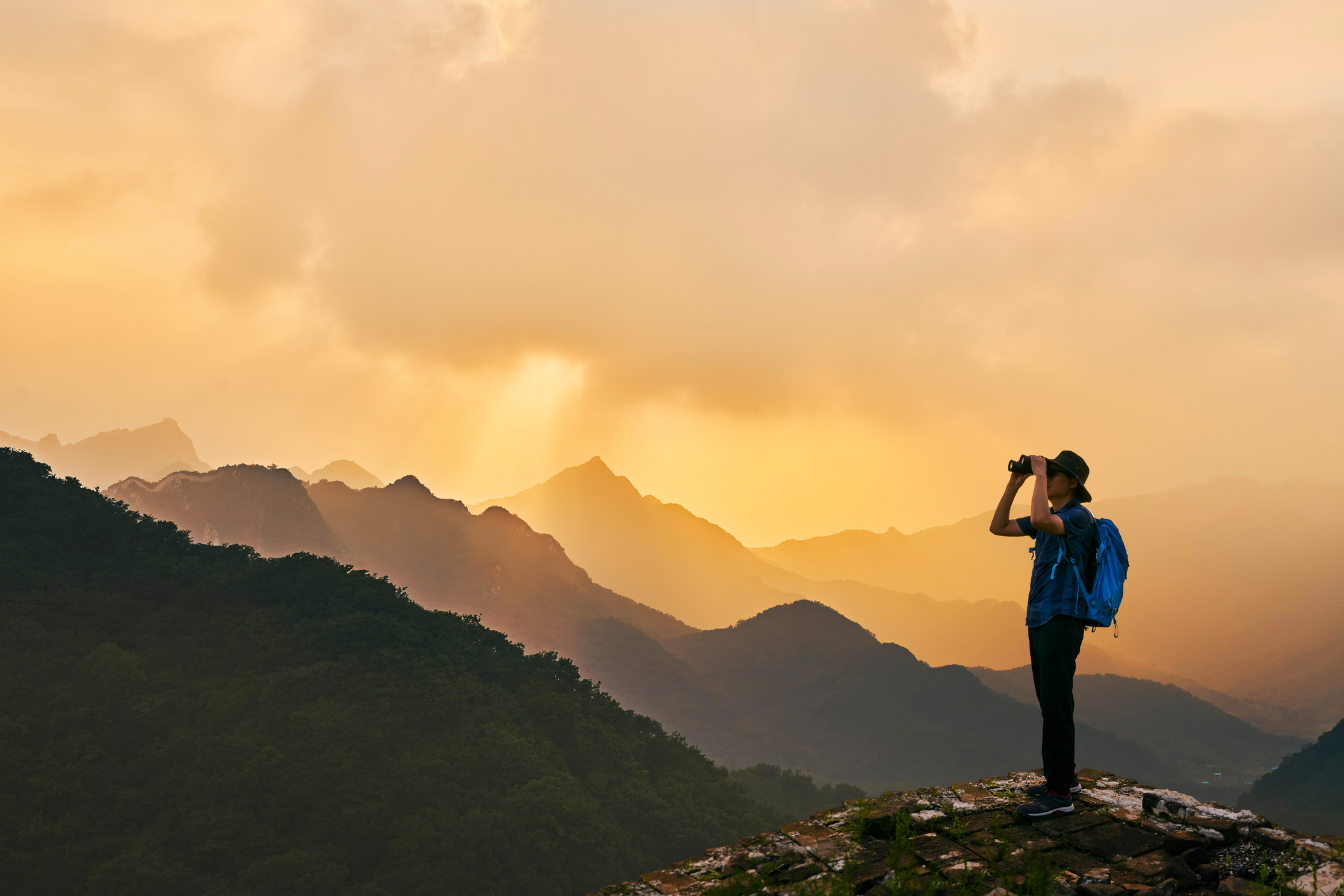 Man Looking Through Binoculars While Standing On Mountain Against Sky During Sunset