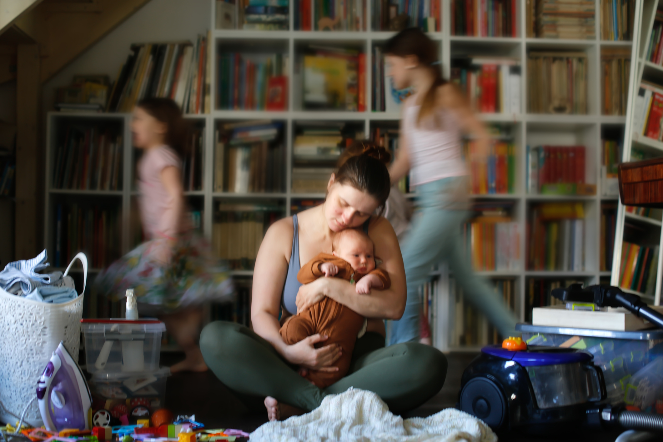 Motherhood Blues: Overwhelmed Mom with Baby, Blurry Running Kids, and the Weight of Household Responsibilities in the Living Room