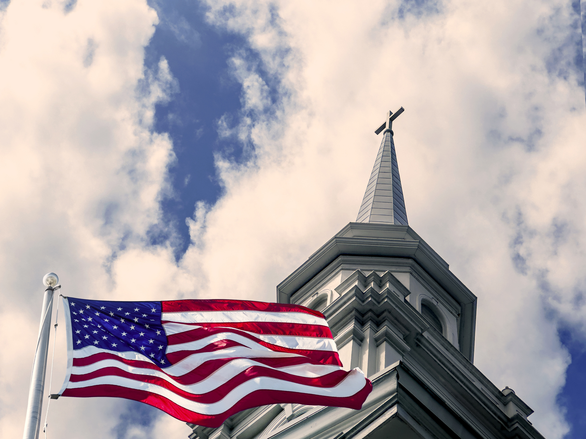 American flag and church rooftop with cross over dramatic sky