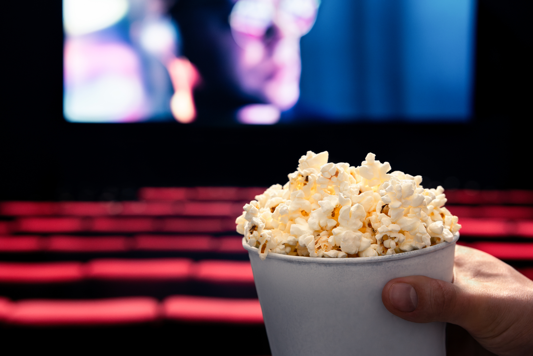 Movies and popcorn. Man holding pop corn box at cinema. Action, thriller or scifi entertainment on screen. Red seats in dark theater. Salty snack in bucket. Spectator pov.