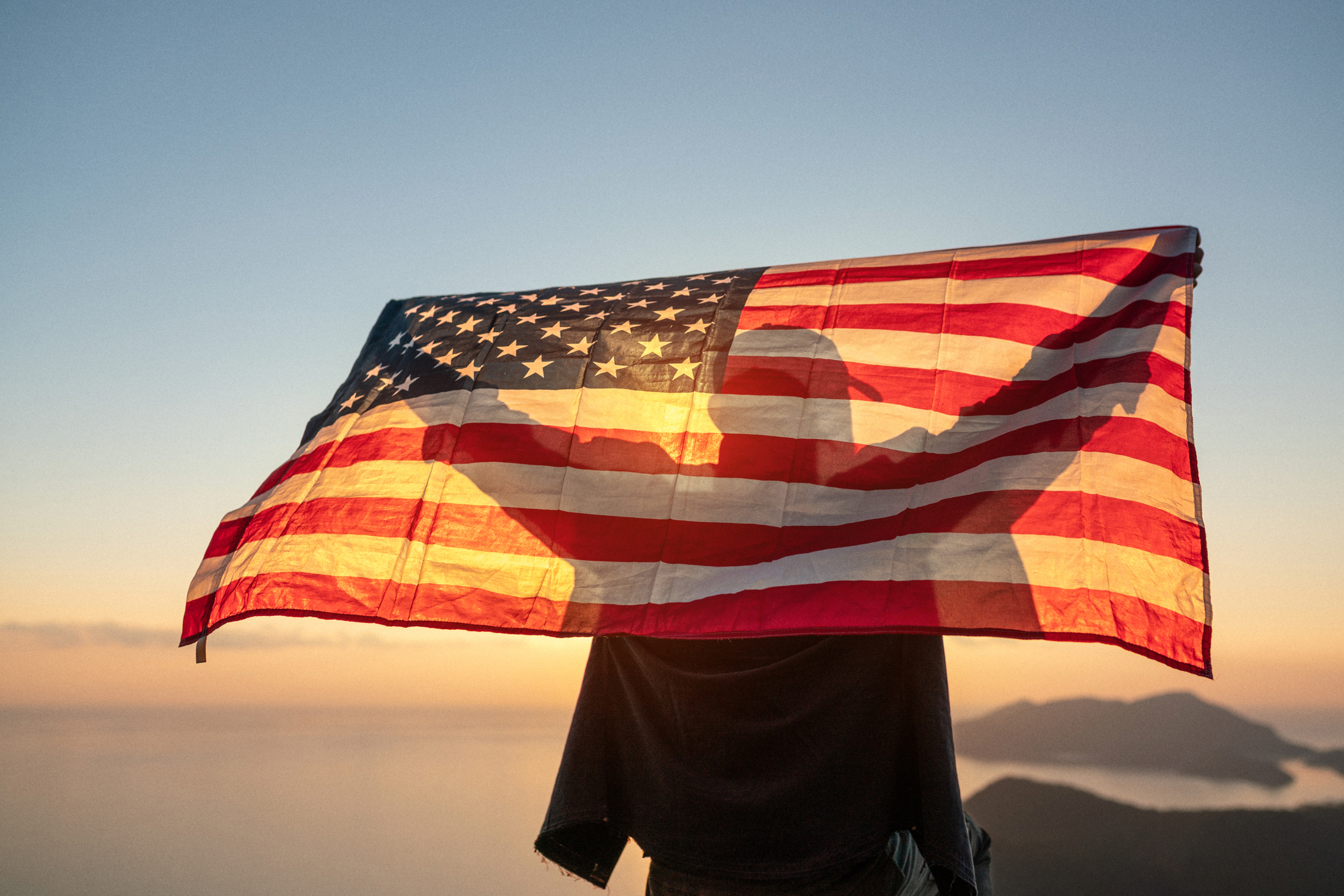 Proud man raising the flag of the United States of America standing on the top of the mountain at sunset