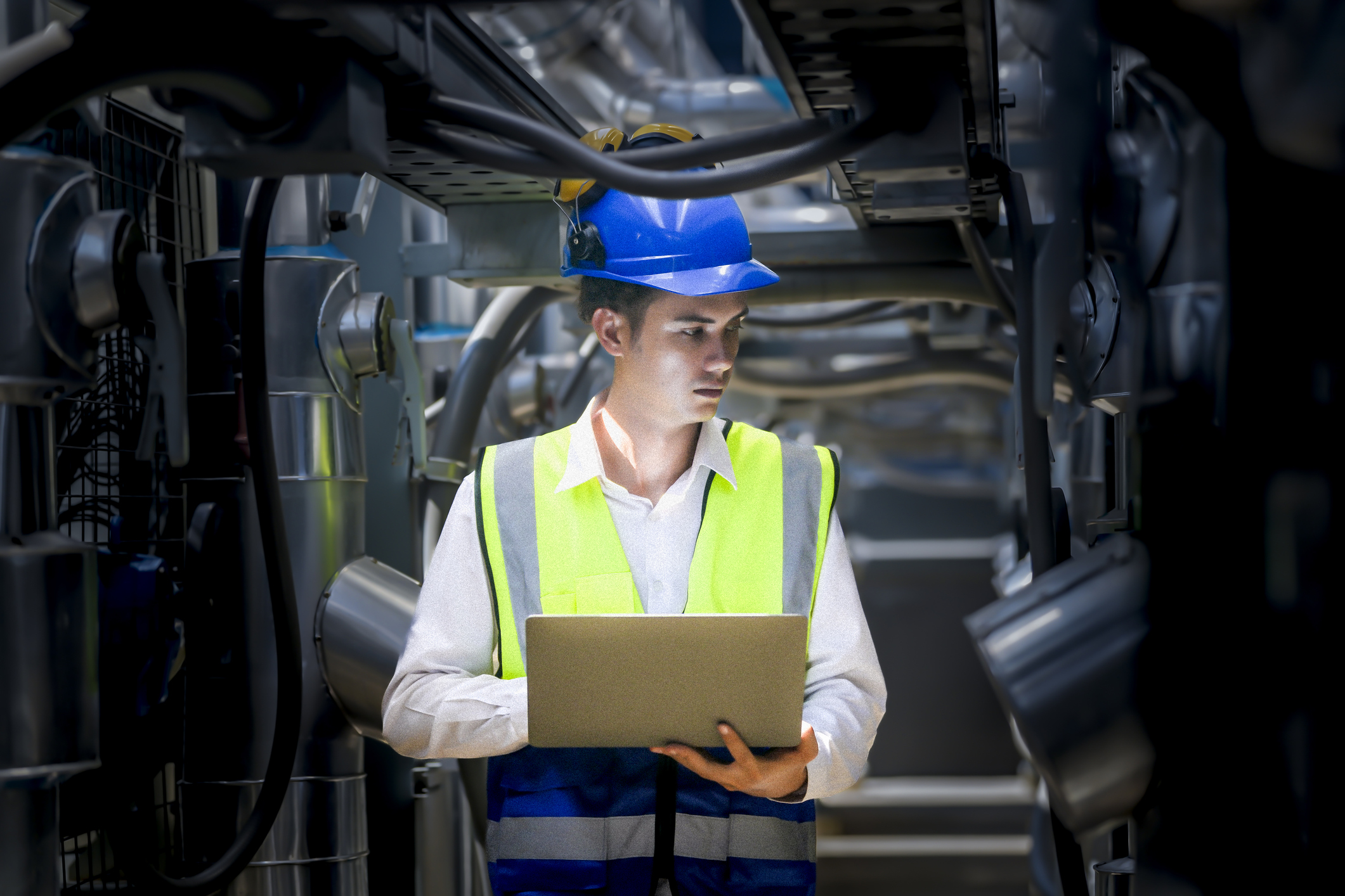 An industrial worker in high-visibility gear and a blue helmet meticulously examines the intricate network of machinery. The engineer is dedicated to maintaining operational efficiency and safety.