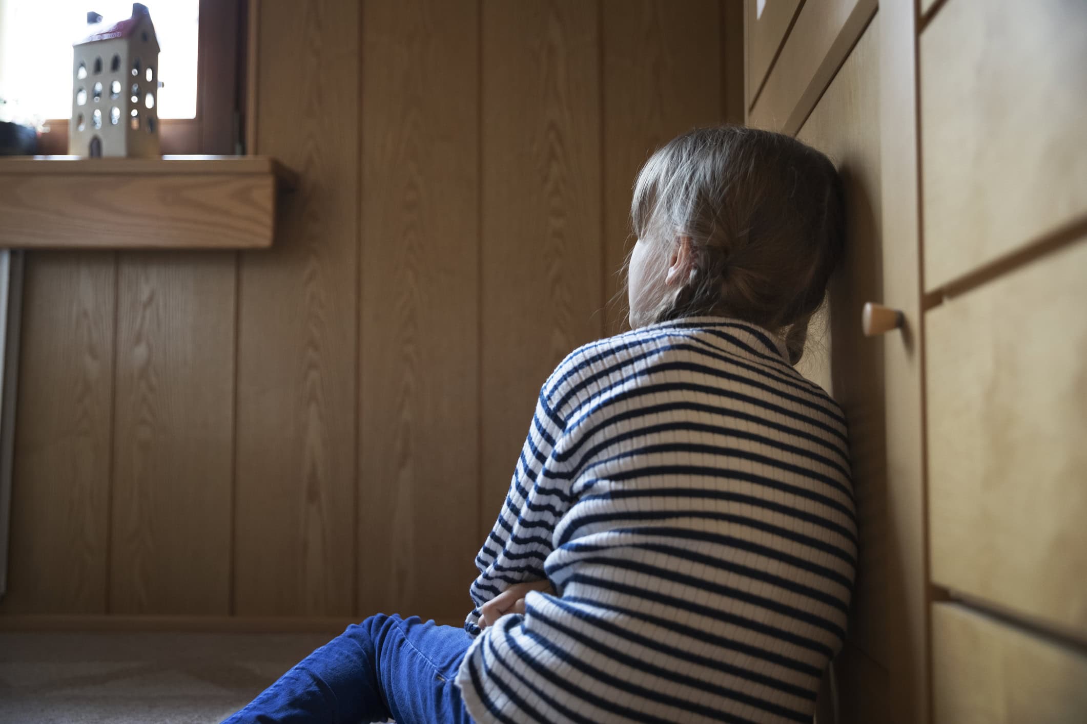 Child sitting on the floor, looking towards a window in contemplation whilst leaning against a wooden cabinet in a living room.